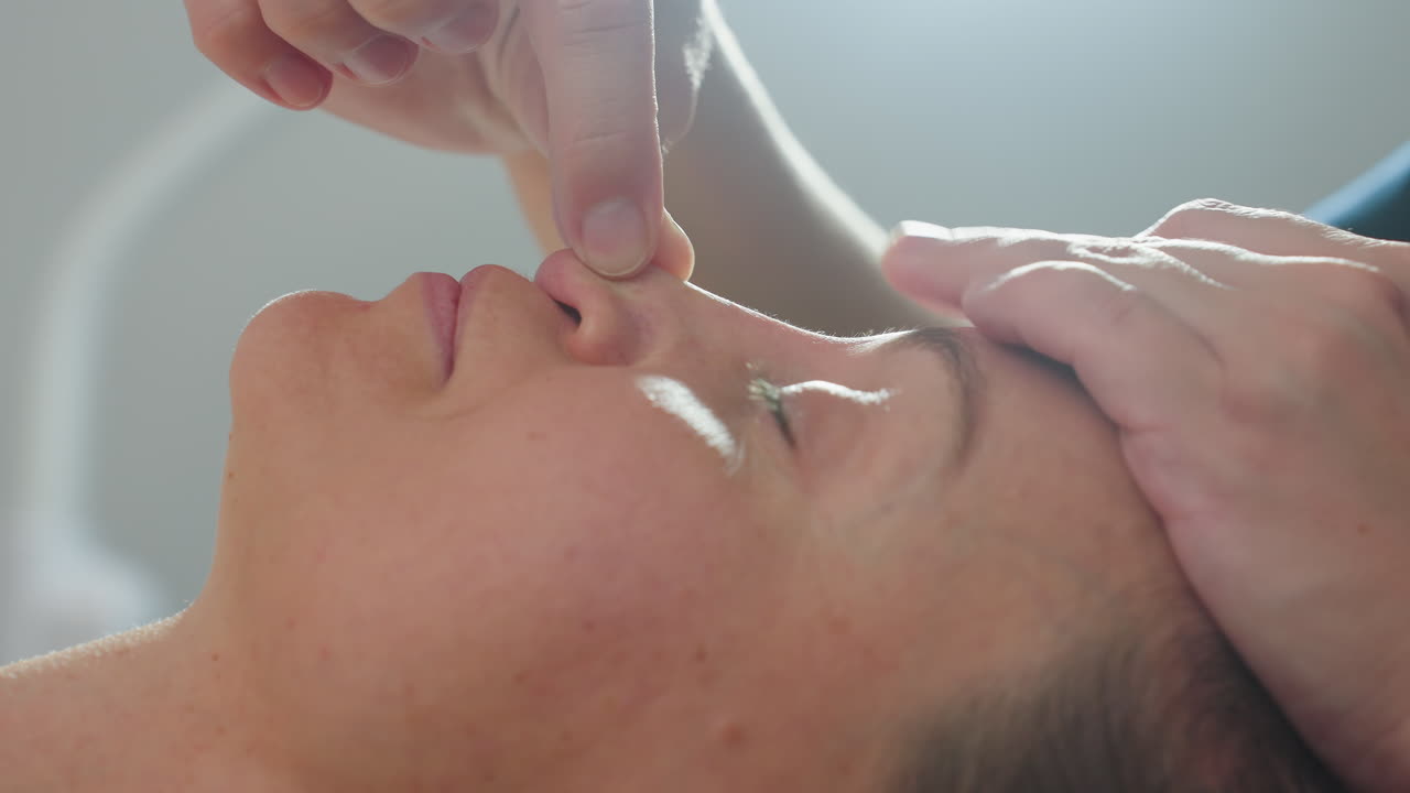 Bodywork expert massages client nose with gentle fingertip movements in calm spa environment, applying smooth pressure along nasal bridge to release tension and ease stress under ambient lighting
