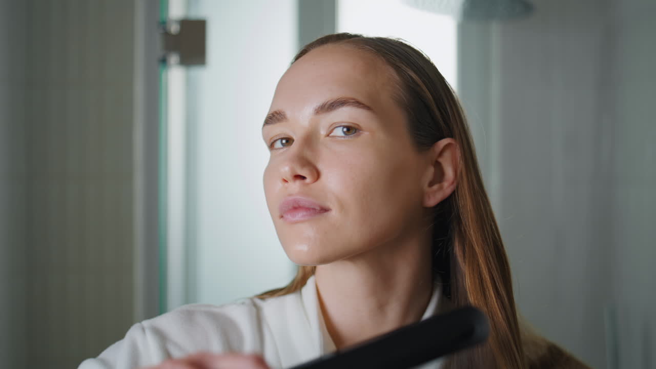 Closeup woman straightening hair in bathroom. Smiling girl getting ready at home