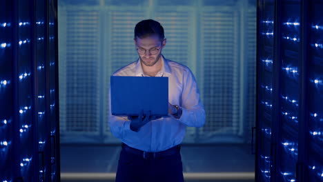 Male-network-engineer-doing-a-system-check-standing-in-the-server-room-with-his-laptop.-At-data-center-men-server-specialists-inspecting-working-system-and-hardware-of-rack-server-computer-cabinets