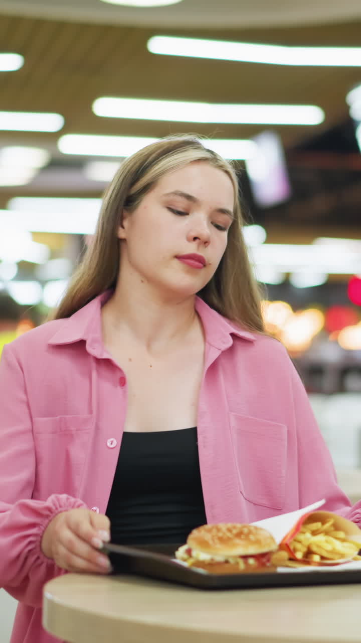 dama de vestido rosa se sienta en el restaurante, papas fritas, hamburguesa, y café en una bandeja, ella alcanza el café con iluminación bokeh y un fondo borroso lleno de gente moviéndose alrededor