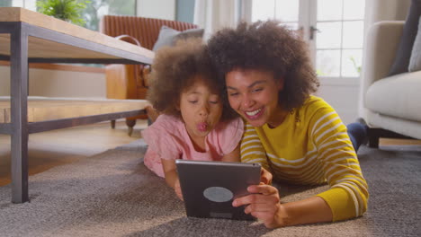 Mother-And-Daughter-Having-Fun-Lying-On-Floor-Pulling-Faces-For-Selfie-On-Digital-Tablet-At-Home