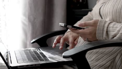 a person multitasks by typing on a laptop keyboard while holding a mobile phone in one hand, highlighting the seamless integration of digital devices into daily routines at home