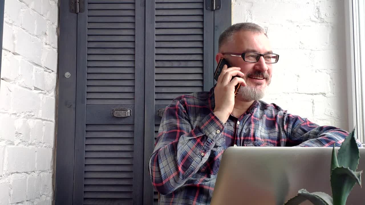 un contable de cabello gris y barba que trabaja en casa detrás de una computadora portátil, haciendo un informe en el telón de fondo de un interior minimalista. él toma una llamada del cliente.