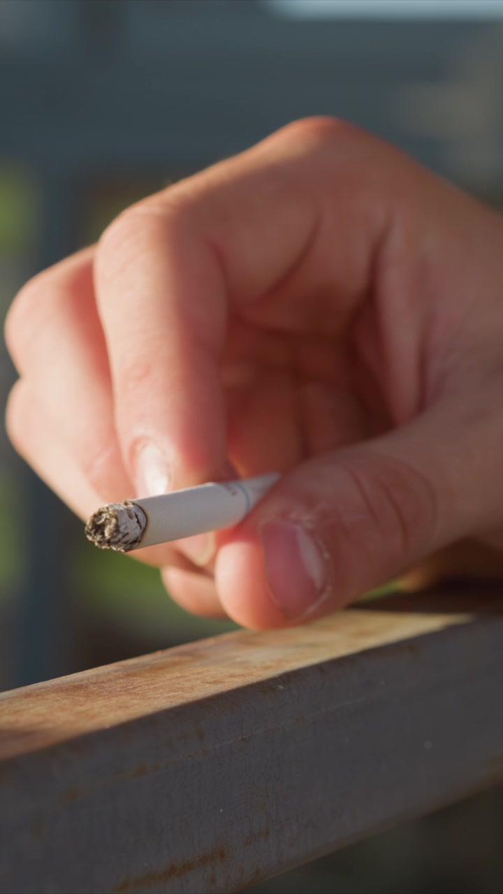 Primer plano de mano sosteniendo un cigarrillo cubierto de ceniza con los dedos sujetando el filtro blanco. La barandilla de metal debajo muestra óxido y detalles de textura bajo una iluminación suave. El fondo está borroso enfatizando