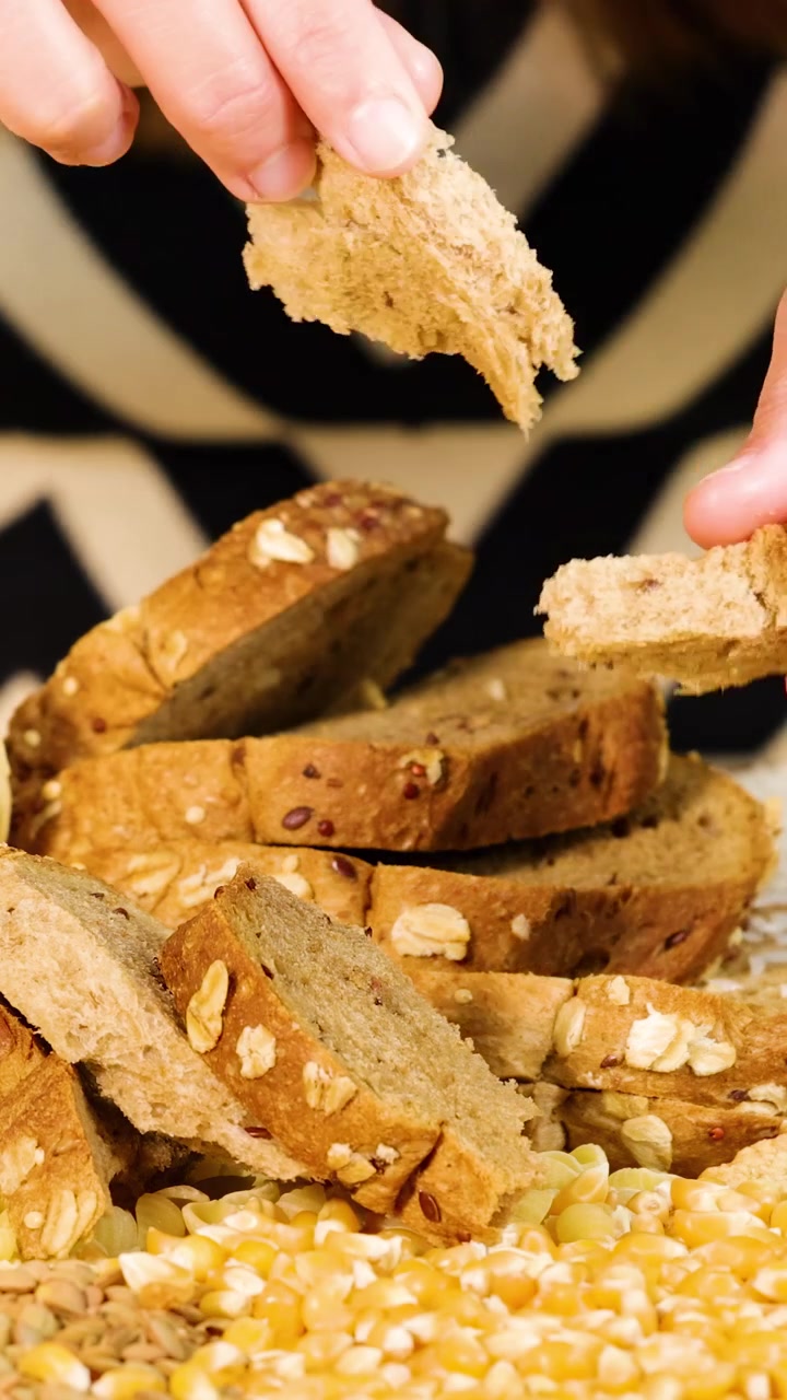 Primo piano di mani che spezzano pane integrale circondato da chicchi. L'illuminazione brillante evidenzia la consistenza e i dettagli