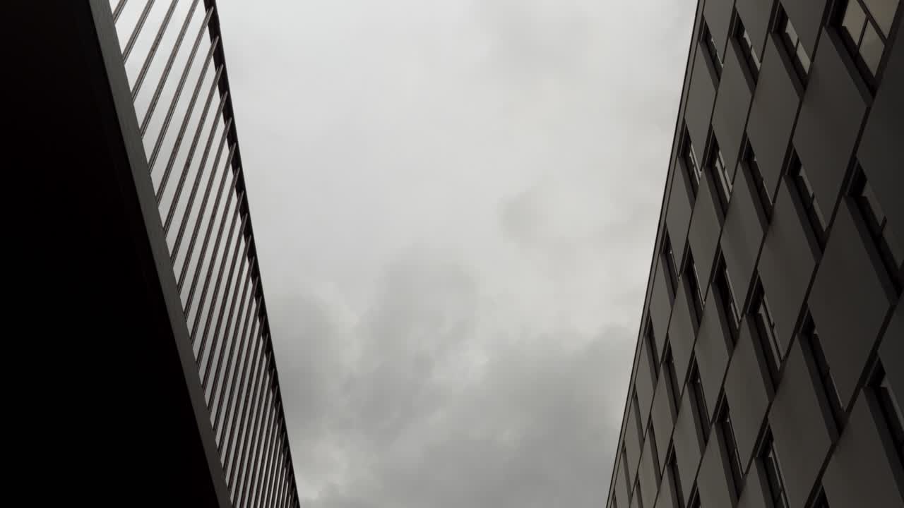 Timelapse looking up between modern buildings as dark clouds race overhead
