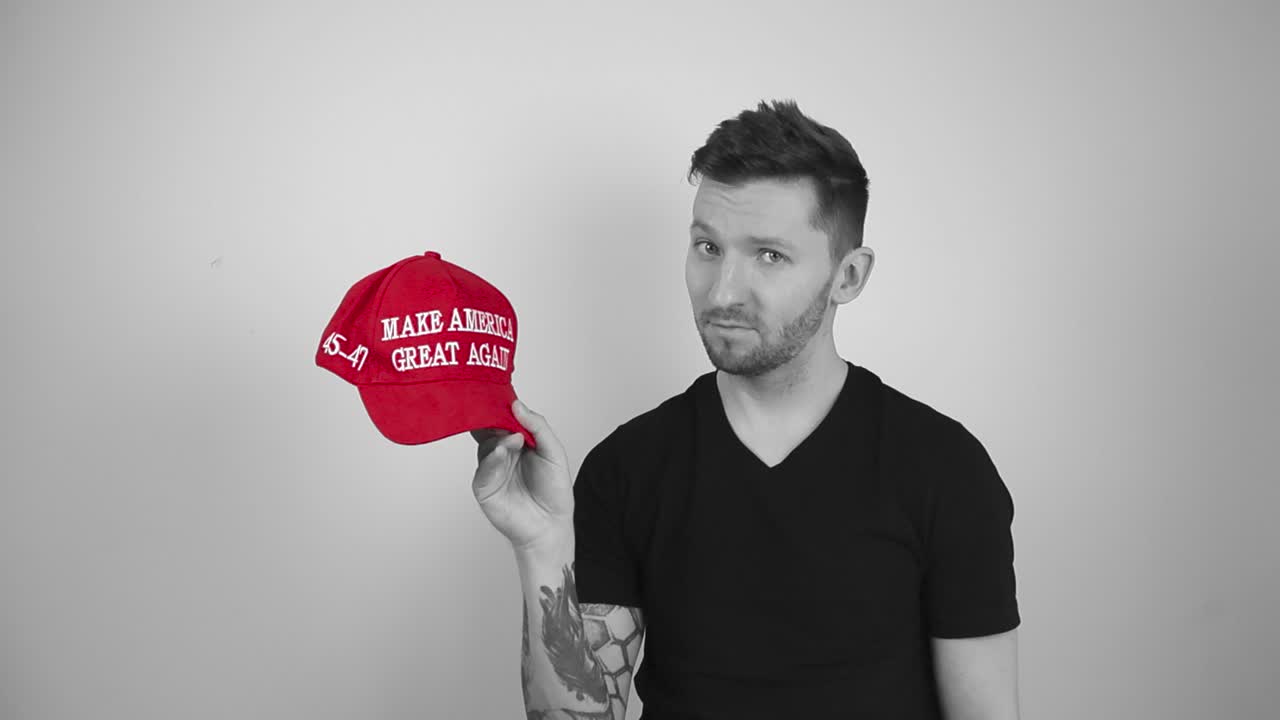 Grayscale footage of a tattooed man lifting up a red vibrant colored MAGA Make America Great Again hat, looking at it and nodding with approval indicating he like the Trump presidential US movement.