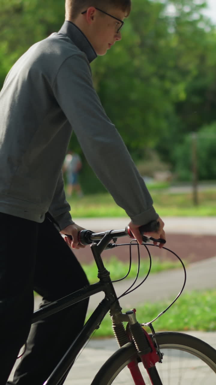 회색 과 검은색 바지를 입은 어린 소년은 느린 동작으로 공원에서 자전거를 타고 있습니다. 배경에는 흐릿한 건물, 나무, 주차된 자동차, 그리고 멀리서 고 있는 누군가가 있습니다.