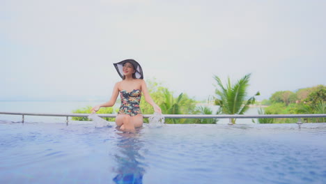 While-sitting-on-the-edge-of-a-resort-swimming-pool,-with-an-ocean-view-in-the-background,-a-young-woman-in-a-bathing-suit-splashes-water-into-the-air