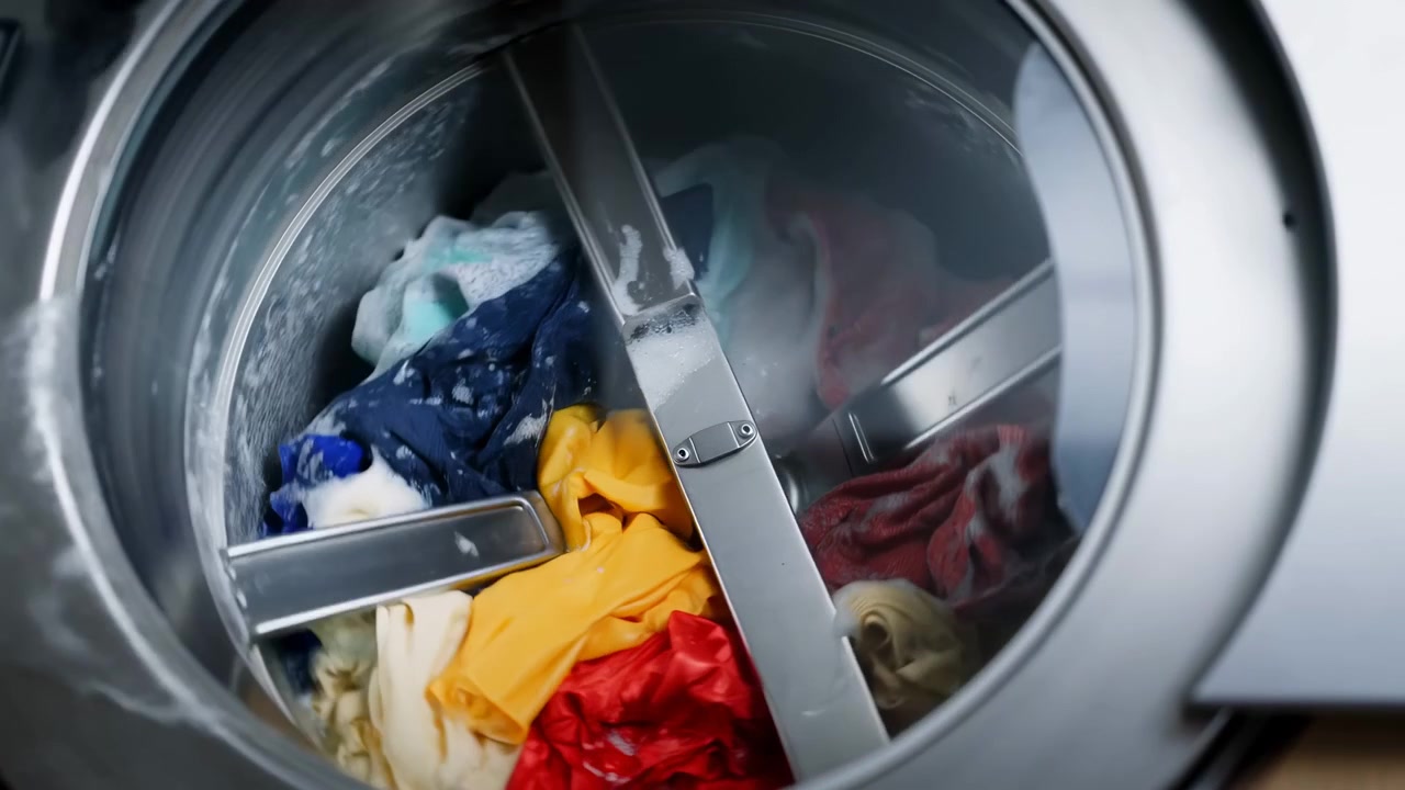 Close-up interior view of a front-loading washing machine with mixed colorful clothes inside a metal drum. Soft indoor light and centered composition suggest household cleaning, laundry care, and everyday domestic routine