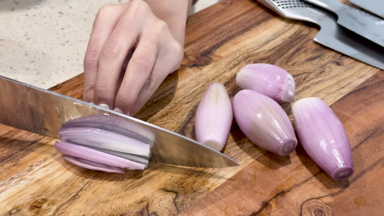A hand uses a chef’s knife to slice fresh shallots on a wooden cutting board under bright, even lighting, with close-up camera framing