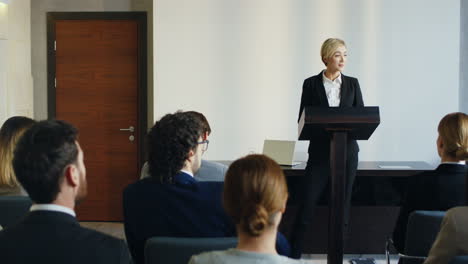 Caucasian-businesswoman-speaker-on-a-podium-at-a-conference-and-interacting-with-people-in-the-hall-during-her-speech