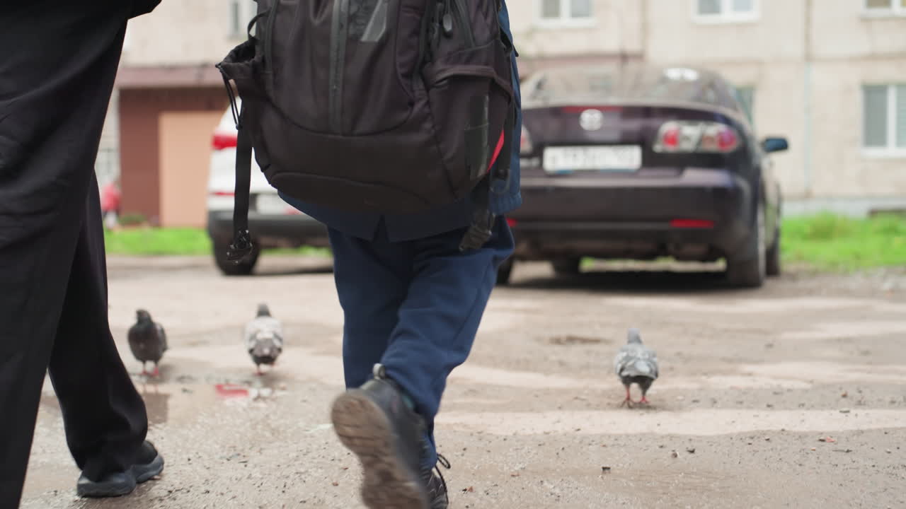 Bambino che cammina con lo zaino e un genitore su un marciapiede urbano, piccioni nelle vicinanze, auto parcheggiate, stivali che schizzano piccole pozzanghere, jeans e giacca, passo costante mentre si prepara per il tragitto scolastico, routine mattutina