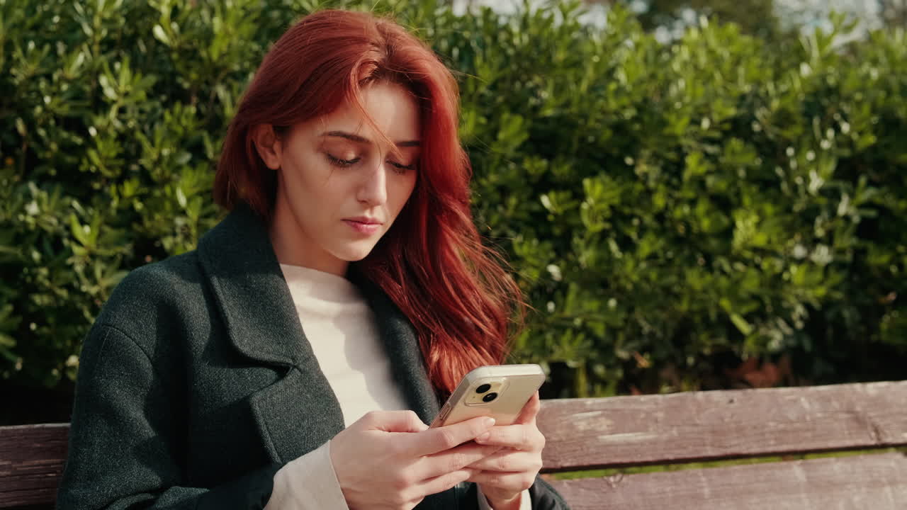 Giovane donna dai capelli rossi che usa lo smartphone su una panchina del parco