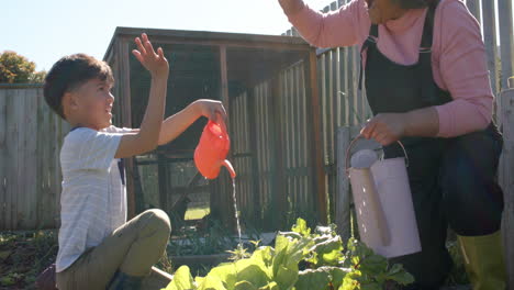 Abuela-Birracial-Mayor-Y-Nieto-Regando-Plantas-En-Un-Jardín-Soleado,-Cámara-Lenta