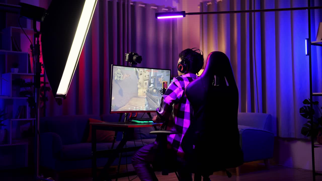 vista trasera de un streamer asiático con dolor de espalda mientras juega un juego a través de la red en una computadora personal. videojuego de transmisión en vivo, escritorio iluminado por luz de tira led rgb
