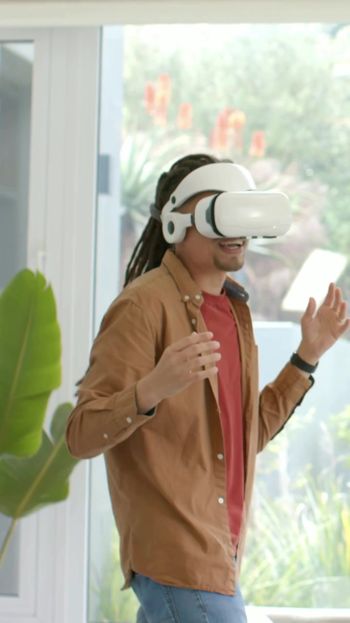 Video verticale: Due amici che indossano visori VR bianchi nel soggiorno, esplorando vicino alla finestra del giardino.