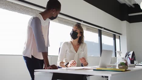 two diverse female colleagues wearing face masks looking at laptop and discussing in office
