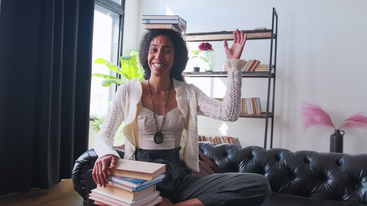 joven atractiva mujer étnica afroamericana con libros en la cabeza se sienta en el sofá