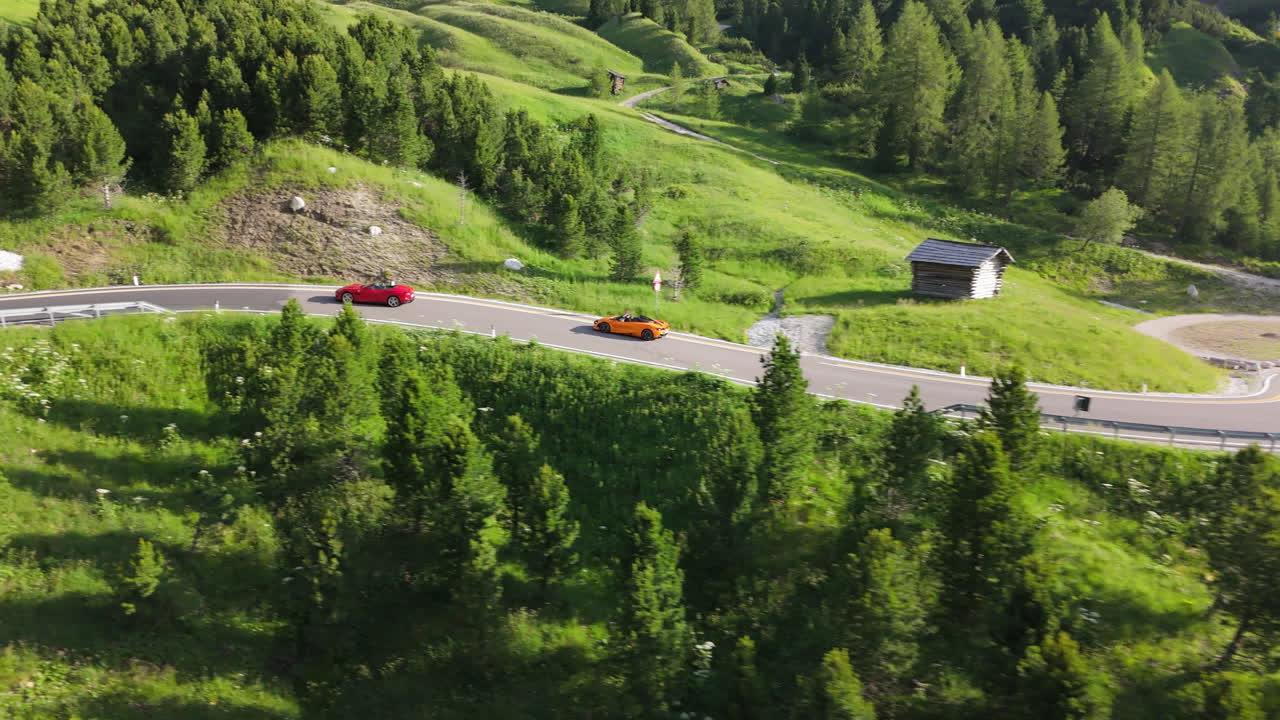 Two sports cars drive through alpine mountains on a winding road during a bright summer morning