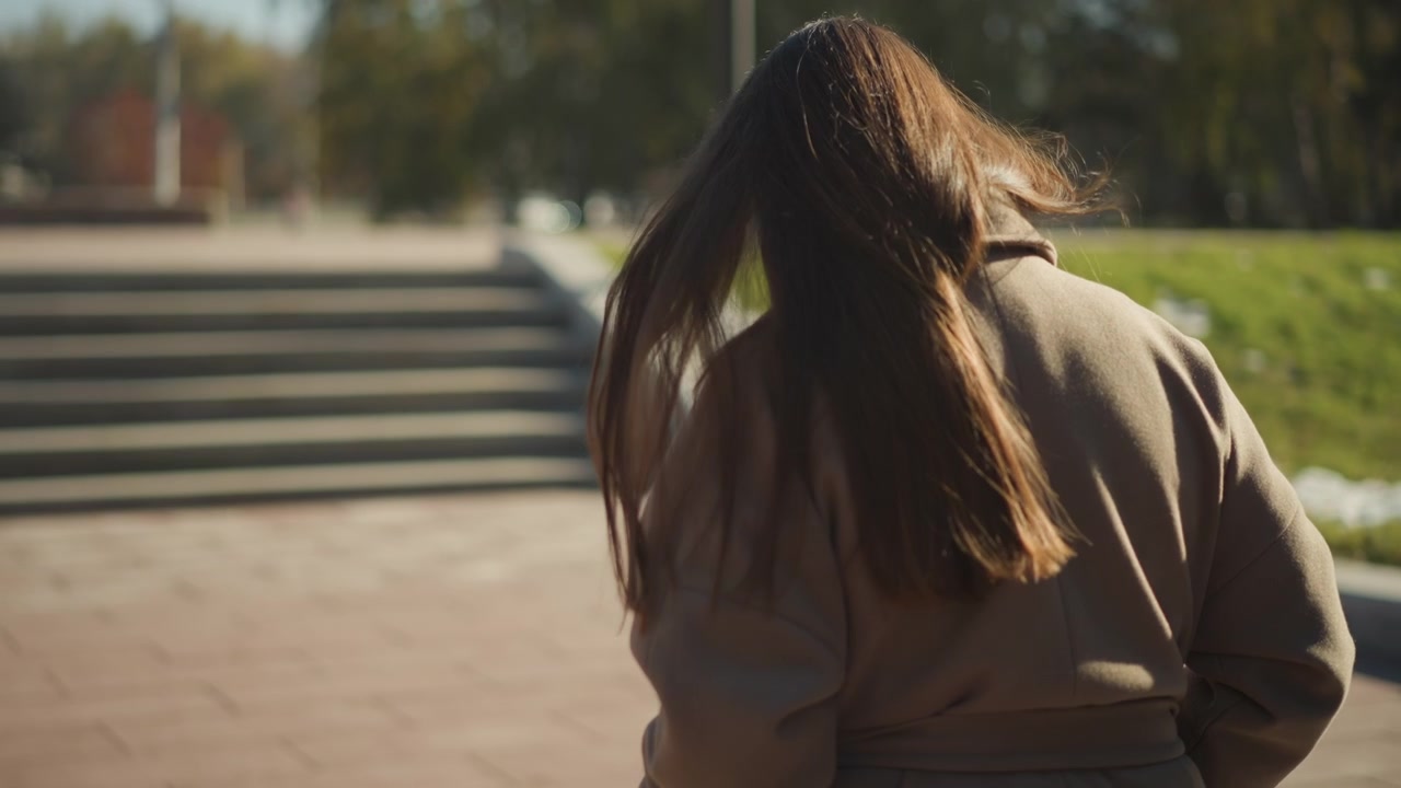 Close up of woman with long hair standing outdoors facing away from camera near concrete steps and green grass. Her brown coat moves slightly in soft lighting suggesting gentle wind