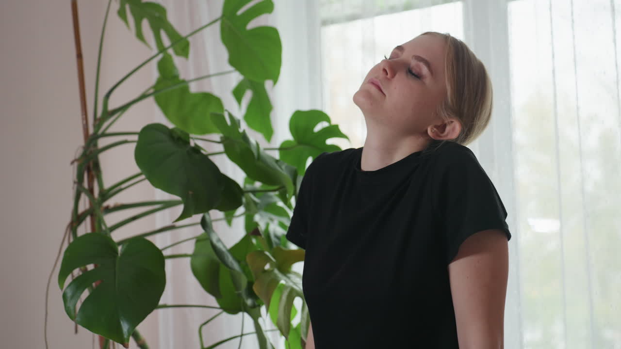 Massage therapist in black shirt gently turning neck for relaxation and comfort while standing beside large indoor plant with natural daylight streaming through sheer curtain in peaceful setting