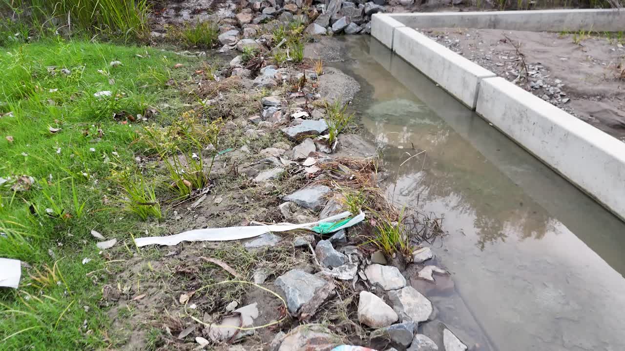 Camera moves along polluted riverbank, highlighting plastic waste, rubbish, and environmental degradation in daylight