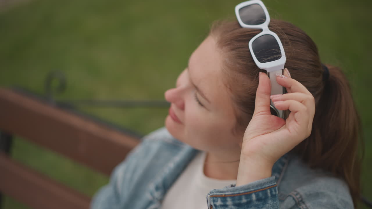 Donna caucasica su una panchina con occhiali da sole, giacca di jeans e coda di cavallo, sotto una luce naturale soffusa. Un sorriso sottile mentre si sistema gli occhiali, con uno sfondo di parco all'aperto. Ritratto ravvicinato di lifestyle, atmosfera estiva rilassata.