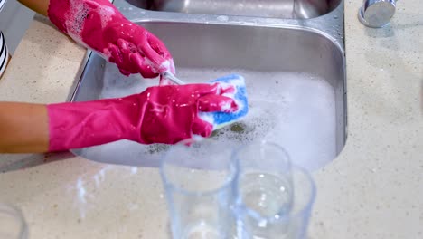 hands in pink gloves wash glasses in a kitchen sink with soapy water