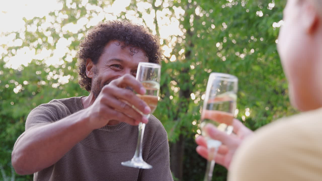 Over The Shoulder Shot Of Mature Couple Celebrating With Champagne Sitting At Table In Garden
