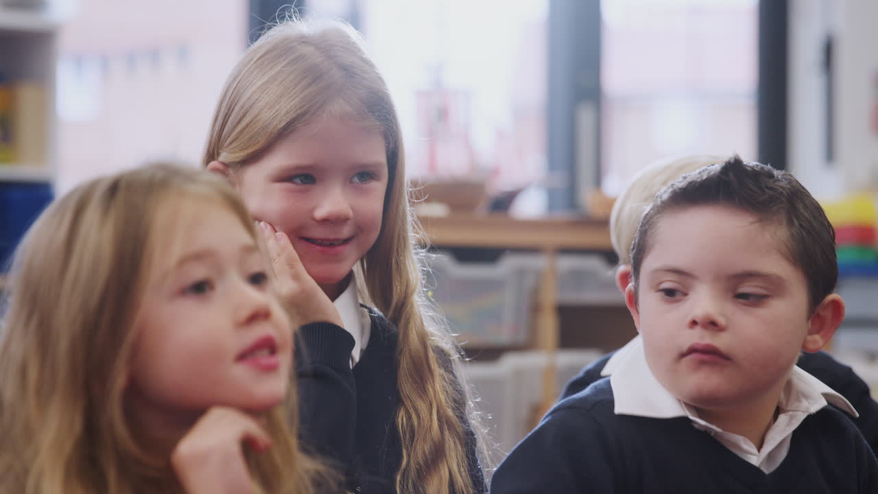 Four primary school kids sitting in class listening and raising hands to answer, close up, backlit