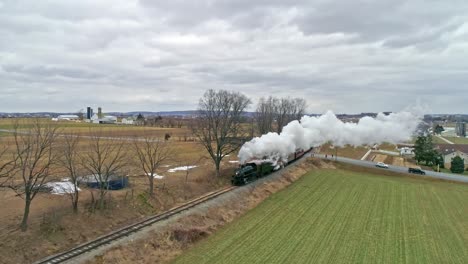 una vista aérea de un tren de vapor con un dron que sigue a un lado soplando humo y vapor viajando por el campo en un día de invierno