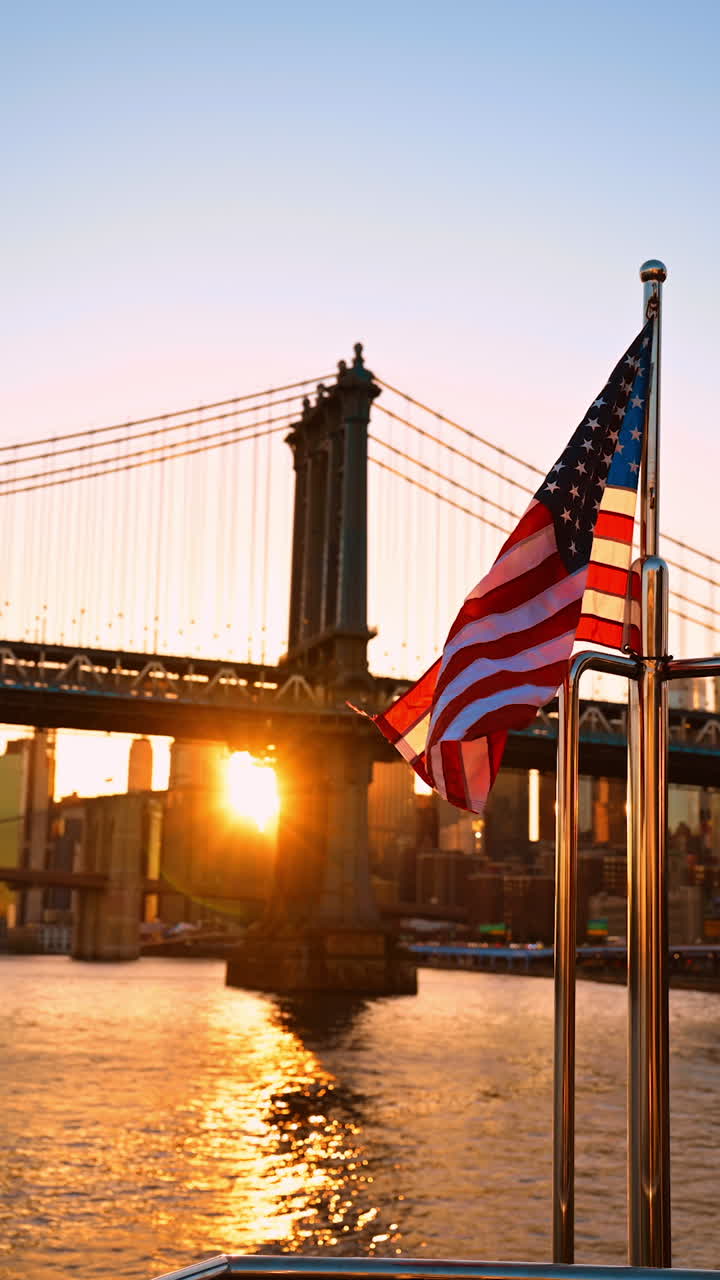 American flag gently flaps in the wind. Bright setting sun dazzles the view among the buildings of New York and the Manhattan Bridge. Vertical video