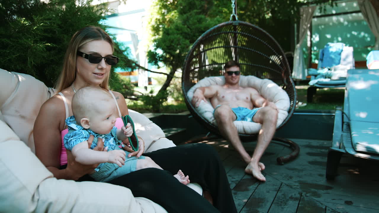 Caucasian family with newborn resting together outdoors in summer. Woman holding a baby and man sunbathing at backdrop.