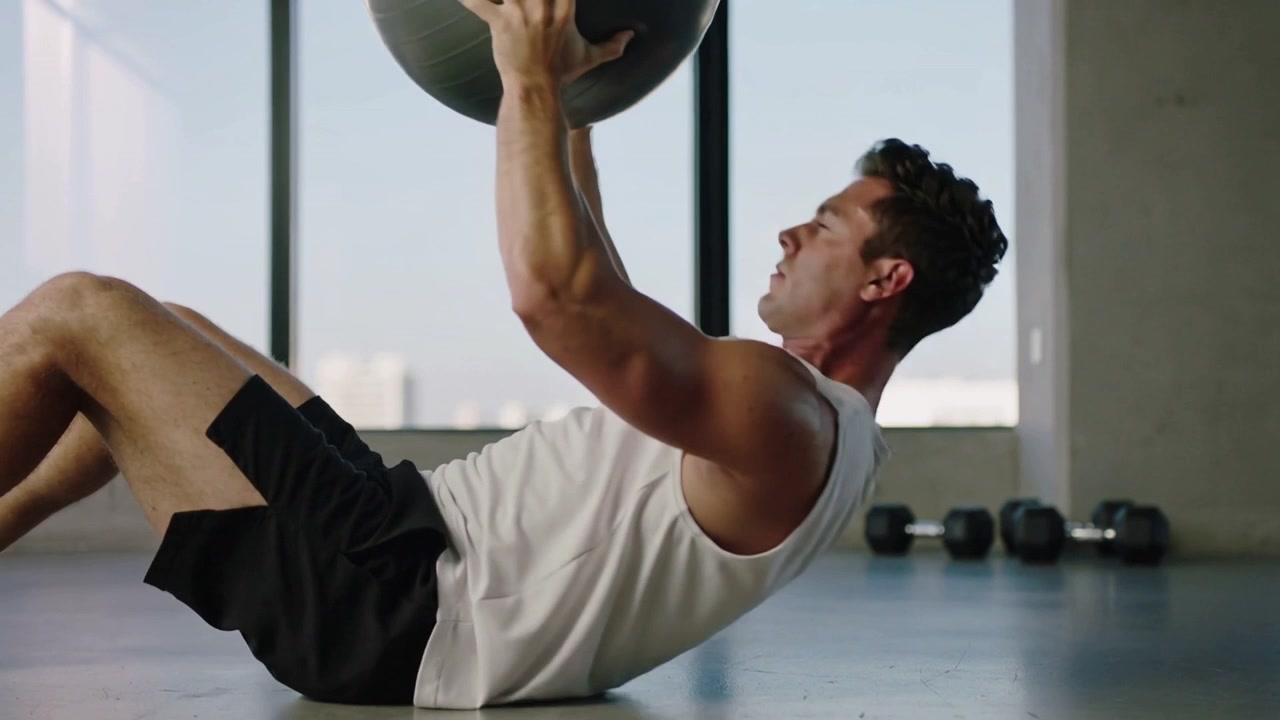 Doing medicine ball crunches, man in white tank, black shorts from back in studio for core