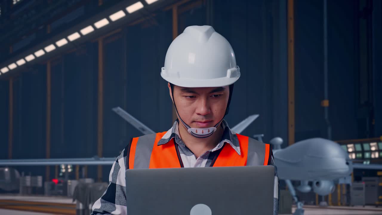 Close Up Of Asian Male Engineer With Safety Helmet Working On A Laptop While Standing, Modern Military Drone in Maintenance Hangar with Industrial Lighting