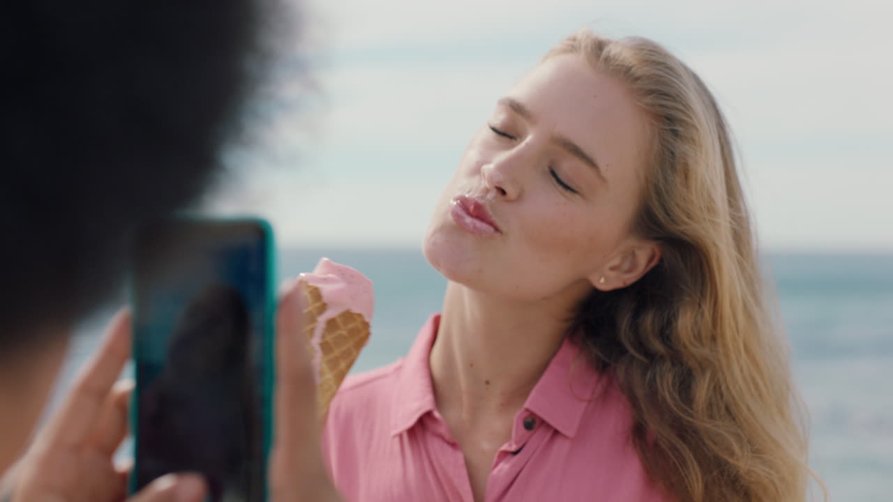 hermosa mujer rubia con afro comiendo helado en la playa posando para una amiga tomando fotos usando un teléfono inteligente amigas compartiendo un divertido día de verano en las redes sociales 4k