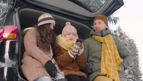 unteransicht eines vaters, einer mutter und einer tochter, die im kofferraum eines autos sitzen, während sie in einem verschneiten wald auf ein smartphone schauen