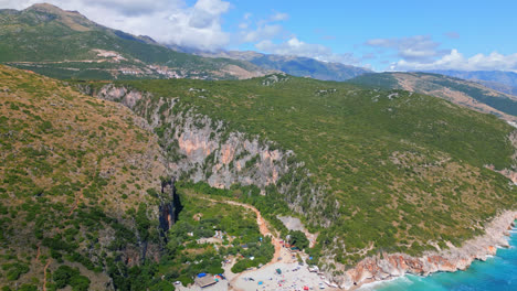 aerial drone backward moving shot over tourists thronging along gjipe beach with canyon in the background in dhermi, albania on a cloudy day