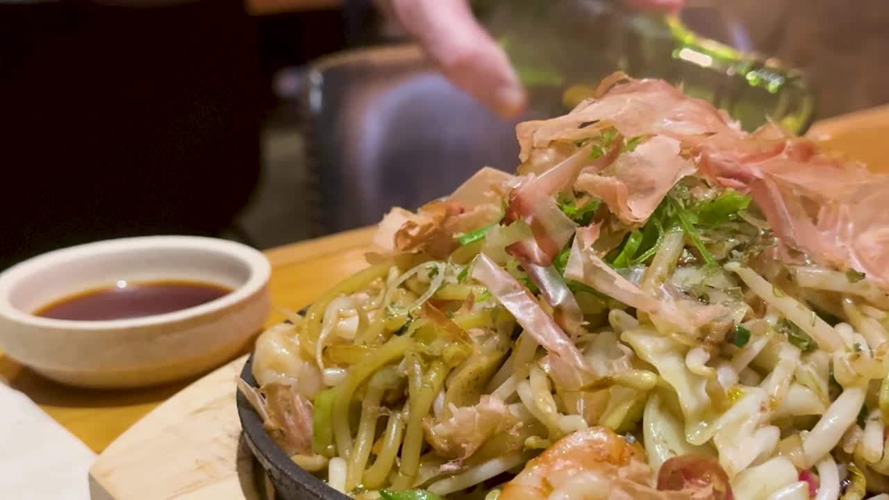 Un piatto fumante di noodles giapponesi saltati in padella, guarnito con fiocchi di bonito danzanti ed erbe fresche.