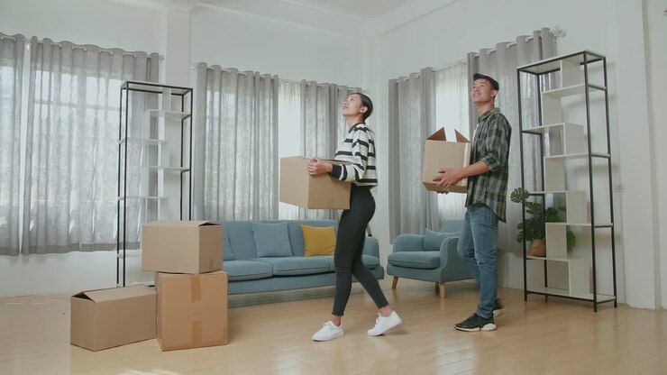 Smiling Young Asian Couple Carrying Cardboard Boxes With Stuff Into A New House Then Looking Around. They Move Into A New House Together