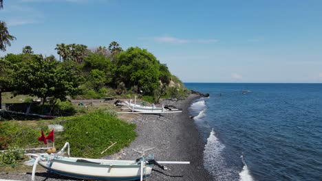 Traditionelle-Balinesische-Fischerkanus-Verstreut-An-Einem-Schwarzen-Vulkanischen-Kiesstrand-In-Der-Nähe-Eines-Kleinen-Dorfes