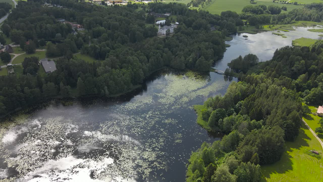 Aerial drone view over scenic Alatskivi Castle lake on radiant summer day. Tranquil water with lilies reflects the sky and sunlight, surrounded by Estonia countryside greenery and manor through trees