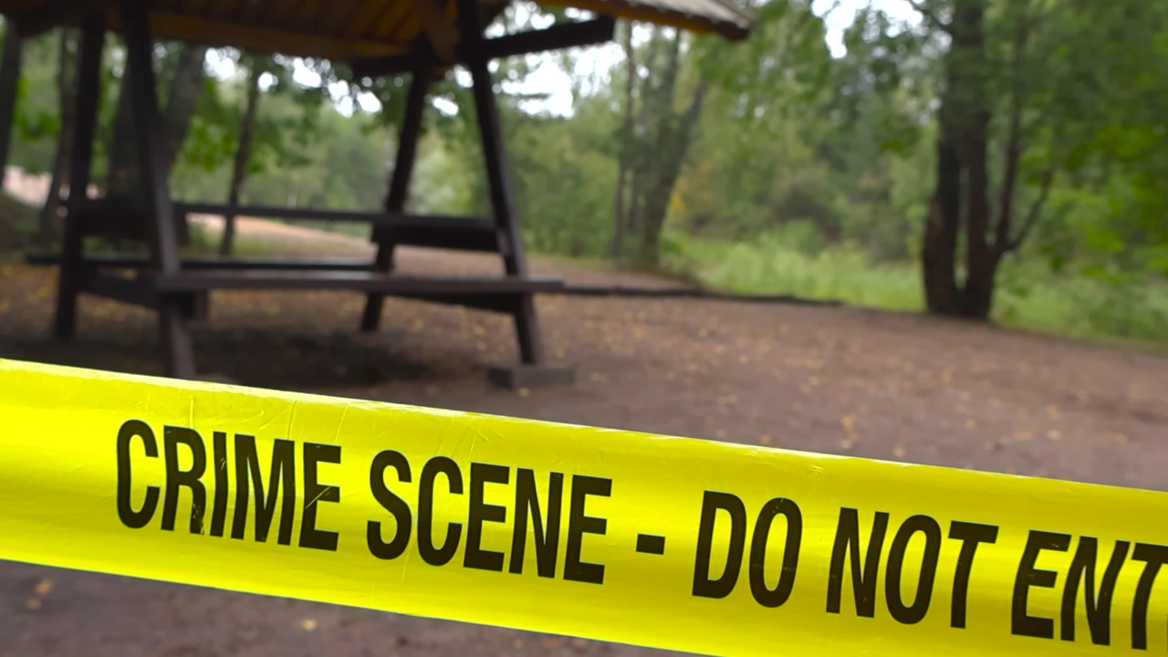 Close up view moving along crime scene tape in focus stretched across blurred forest camping area with picnic table background on moody, overcast summer day. Recreation spot marked investigation site