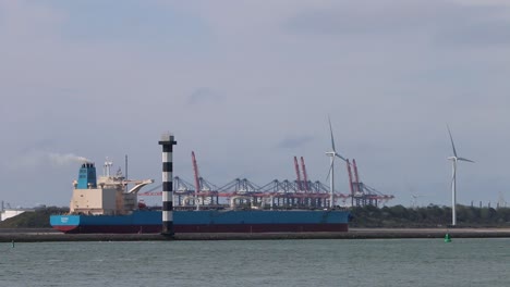 barco comercial que sale de los muelles, con grúas y turbinas eólicas al fondo