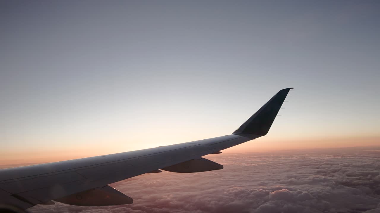 View out window of commercial airline plane wing with soft orange and magenta hues at sunset with clouds below