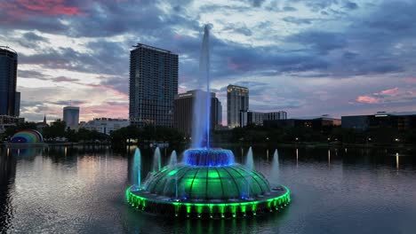 aerial orbit shot of lake eola with fountain in colorful lights. twilight scene with skyscraper of downtown in background. wide shot. financial district of orlando florida.