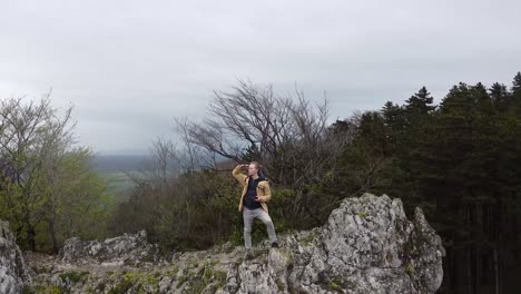 Reisender-Mit-Rucksack-Erreicht-Die-Spitze-Einer-Steilen-Bergklippe-Mit-Flachen,-Farbigen-Waldhügeln-Und-Blickt-Auf-Die-Landschaft