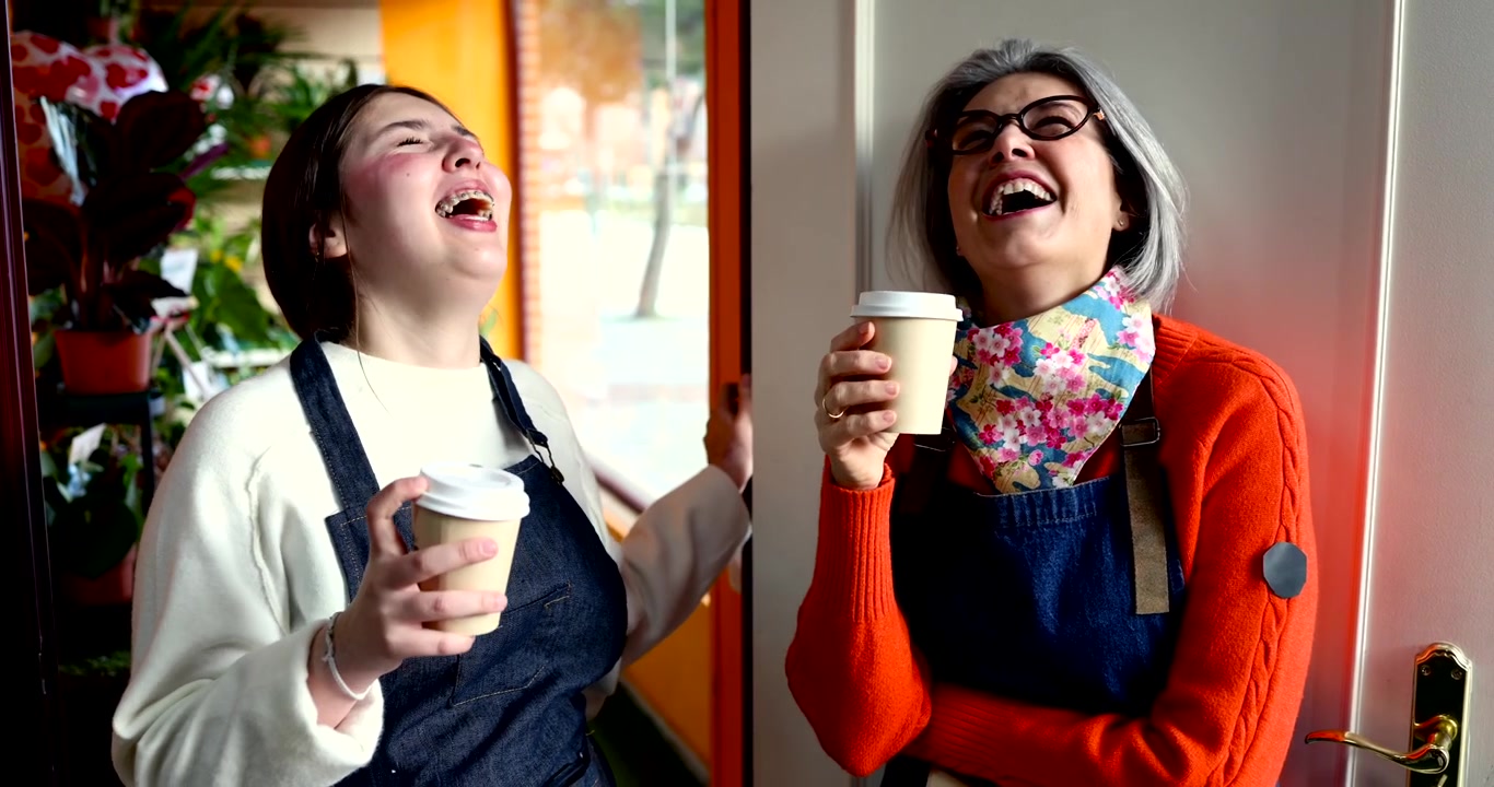 Two Women Wearing Aprons Laughing and Drinking Coffee in a Flower Shop