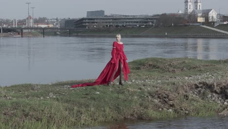 модель, стоящая на небольшом острове в ярко-красном платье с городом на заднем плане